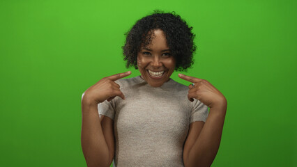 Young african american woman smiling and pointing finger to cheek against green studio backdrop; confidence.
