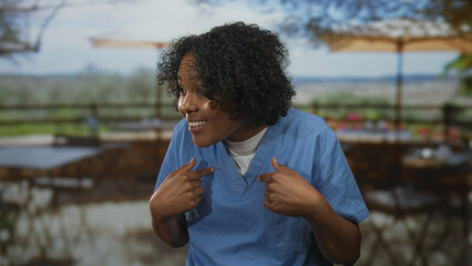 Naklejka premium Smiling woman pointing her thumbs to her chest on a restaurant terrace overlooking landscape; inner pride.