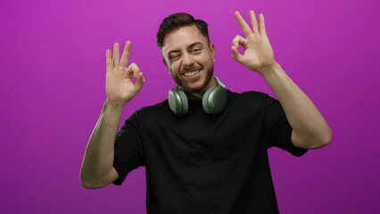 Fototapeta premium Young man with headphones smiling and making an ok hand gesture against a vibrant pink background, exuding confidence and positivity.