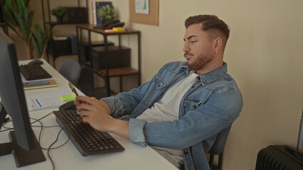 Young man with beard using smartphone in modern office workspace with computer and shelves, displaying focus and concentration