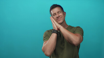 Man with hands to cheek for sleep wearing olive t shirt in turquoise studio with closed eyes and...