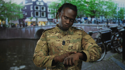 Man soldier in camouflage points finger upward on an amsterdam canal street while checking wristwatch and wearing dog tags; duty vigilance resolve.