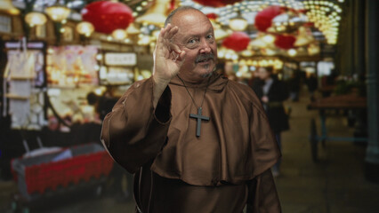 Man monk makes ok hand signs showing cross necklace in a decorated building market with stalls and hanging lights; faith celebration cheerful.