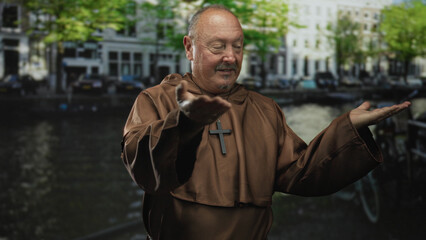 Man in brown robe extends hand showing cross necklace on street beside canal and building facades;...