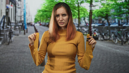 Fototapeta premium Woman holding a pair of yellow socks at shoulder height, frowning with knitted eyebrows and slightly hunched posture on a bike lined cobblestone street; frustration.