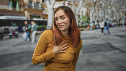 Fototapeta premium Woman with red hair places hand on chest in a sunlit street, smiling in a mustard sweater with a bus and tree lined avenue behind her; gratitude warmth.