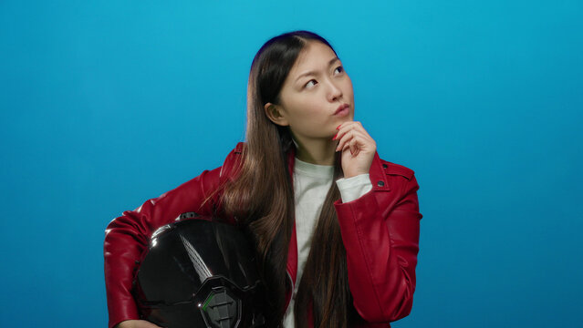 Woman in red jacket pondering holding helmet against blue background with thoughtful expression implying decision-making setting.