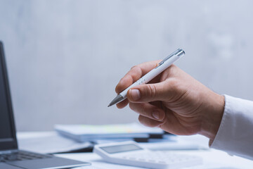 Male hand with pen reviewing printed reports and charts on laptop desk. Professional business, finance, audit and corporate workflow concept with copy space.