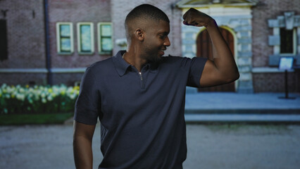 Man flexes bicep with arm bent at elbow, smiling and looking at his raised arm beside a stone building entrance with steps and tulip flowerbed; confidence strength.