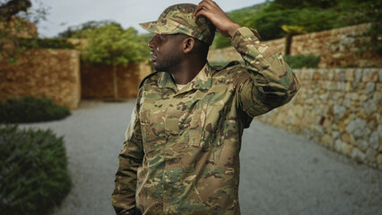 Soldier adjusts cap, hand on head, looking sideways in camouflage uniform on stone street with walled courtyard and shrubs; uncertainty duty.