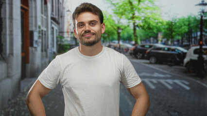 Fototapeta premium Man in white t shirt and beard standing with hands on hips on a city street near parked cars and trees, slight smile with closed eyes; confidence.