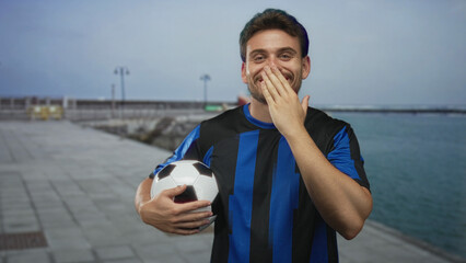 Man holding soccer ball with hand over mouth on street promenade by the sea wearing blue jersey, smiling; joy team spirit.
