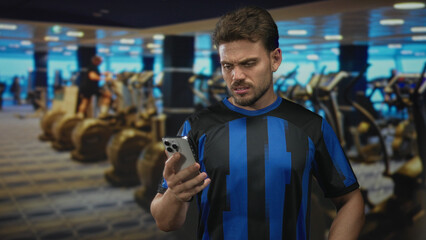 Young hispanic man holding a smartphone, frowning at the screen in a gym building while checking messages; frustration distraction.