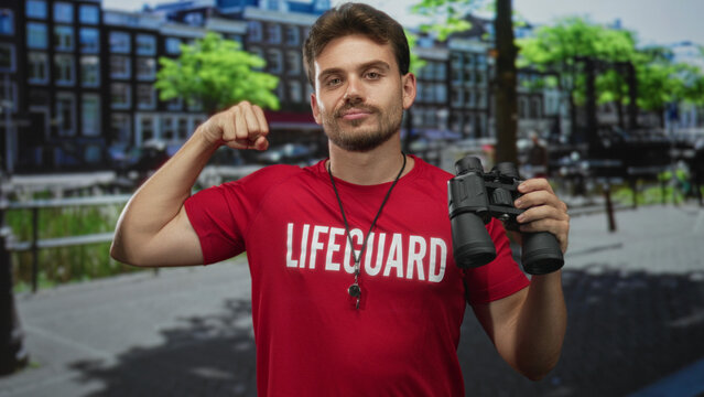 Man lifeguard flexing biceps wearing red shirt and holding binoculars on amsterdam street; confidence rescue readiness.