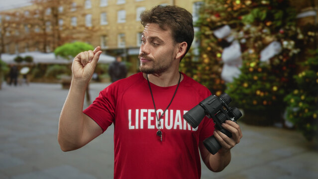 Man lifeguard holding binoculars and pinching fingers near eye on street, whistle visible, red shirt; vigilant duty.
