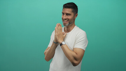 Man pressing palms together in a teal studio, smiling and leaning forward with watch visible;...