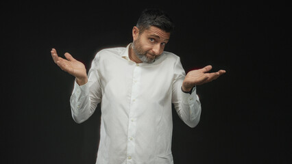 Man shrugging with open hands and raised shoulders in a black studio, casual white shirt and...