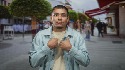Fototapeta premium Young hispanic man holding jacket with hands and thumbs to chest on a busy shopping street lined with shops and trees; uncertainty.