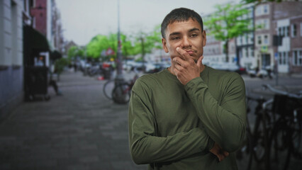Fototapeta premium Man touching chin in green long sleeve shirt on a street with bicycles and buildings; thoughtful reflection.