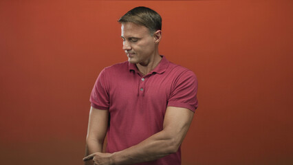 Man pointing with right hand and visible forearm while wearing a red polo shirt against an orange studio backdrop, looking slightly downward; steady confidence.