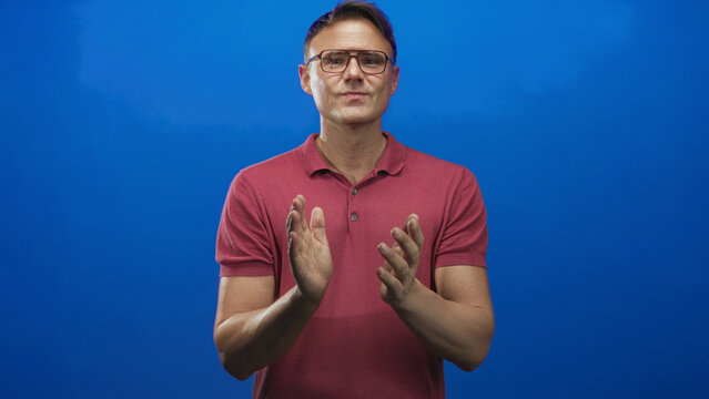 Man clapping hands in a studio with blue wall, wearing glasses and red polo shirt; warm approval encouragement. - Powered by Adobe