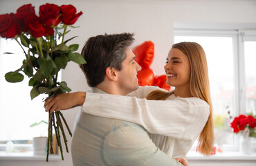 Smiling young couple embracing at home as the man lifts his partner holding a bouquet of red roses during Valentine’s Day celebration.
