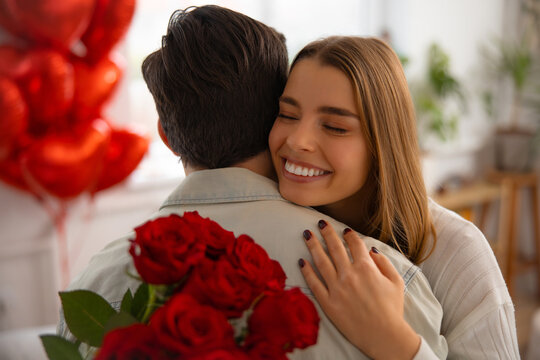Smiling woman hugging her boyfriend after receiving red roses on Valentine&rsquo;s Day at home.
