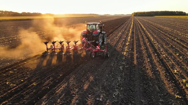 Tractor Plowing Field at Sunset - A red tractor with a plow is seen working a field at sunset, creating a cloud of dust behind it.