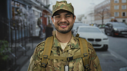 Fototapeta premium Man wearing camouflage uniform with backpack straps and dogtag smiles in urban street setting; pride duty honor.