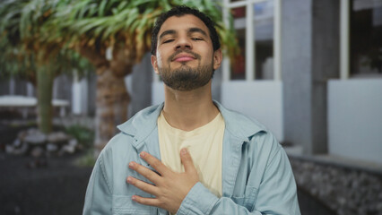 Young hispanic man places hand on chest while standing on a city street by building facade; sincere...