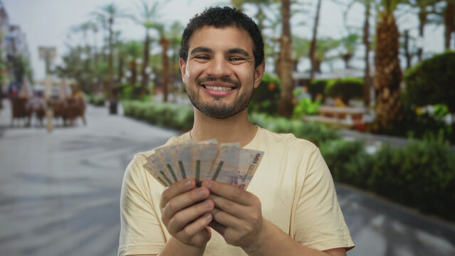 Young hispanic man holds saudi riyal banknotes in hands on street with palm trees and green foliage; prosperity.