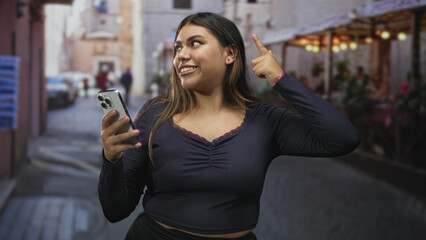 Woman holding smartphone points finger upward on a narrow street lined with cafes; confidence and...