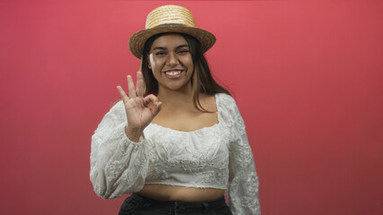 Woman making ok hand gesture, cropped top showing midriff and straw hat in studio; confidence summer gesture.