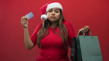Woman holding shopping bags and a creditcard, wearing a santa hat and frowning while holding up the...