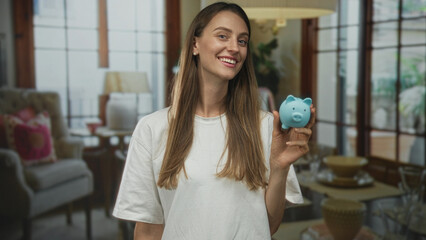 Woman smiling while holding blue piggy bank with hand in building living room near lamp and chair; saving money optimism.