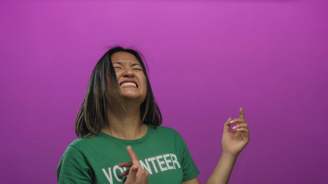 Young chinese woman playfully mimicking guitar playing with enthusiasm, wearing a green volunteer t-shirt against a vibrant pink wall in a joyful expression of creativity. - Powered by Adobe