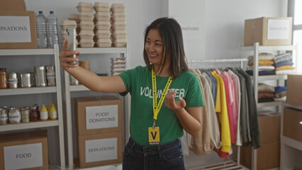 Woman smiling during videocall in donations center surrounded by clothing and food items wearing green volunteer shirt in indoor room setting suggesting charitable activity.