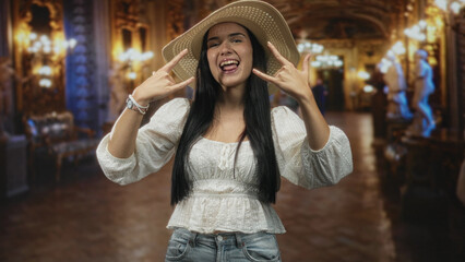 Woman sticking tongue out and flashing rock horns with both hands in ornate historic building lobby, wearing straw hat, white blouse and denim jeans; playful rebellion.