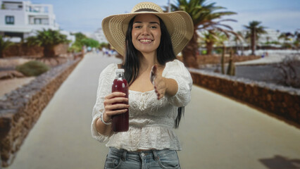 Woman holding a juice bottle with her hand offering it on a street promenade with palm trees;...