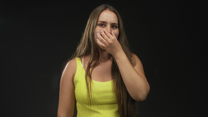 Young woman pinches nose with hand while wearing a yellow tank top in a studio with black backdrop; disgust smell repulsion.