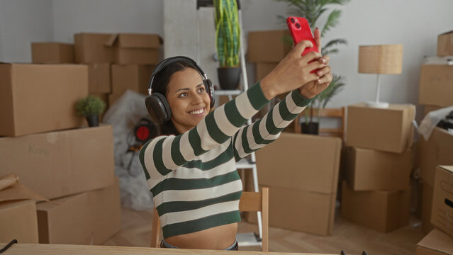 Young woman taking selfie in new home surrounded by moving boxes while wearing headphones and smiling at camera in bright apartment setting.