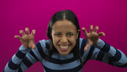 Woman playfully making claw gestures in front of a vibrant pink background wearing a striped sweater and expressing joy with a lively and engaging expression.