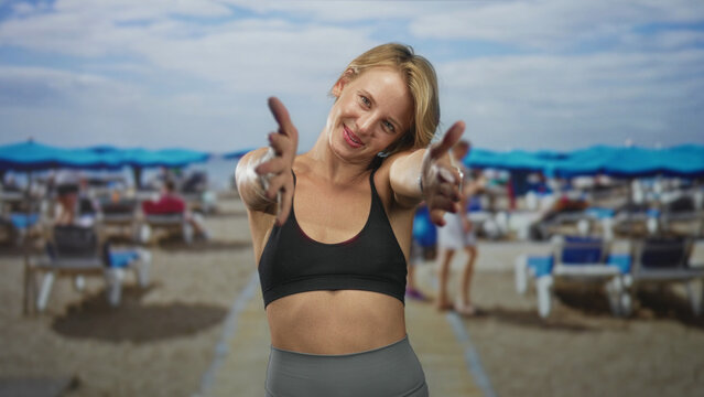 Woman hugging shoulders with crossed arms, smartwatch on wrist, wearing black sports bra and gray leggings on a sandy beach with blue umbrellas and lounge chairs; joyful relaxation. - Powered by Adobe