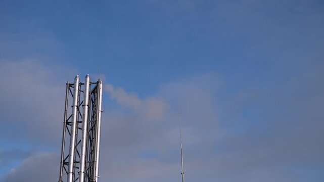 industrial steel tower against blue sky, tall metal lattice mast with antenna, calm daylight and scattered clouds, urban infrastructure silhouette, maintenance ladder and platform visible,