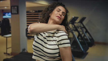 Woman in striped shirt touches shoulder and collarbone inside a gym building with closed eyes and...