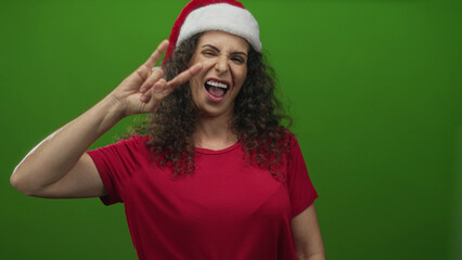 Woman wearing santa hat and red shirt makes rock horns gesture with hand against vivid green studio...