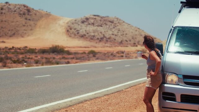Young woman leans against a white van while sipping coffee. She enjoys a quiet moment on an empty desert road under a clear sky.