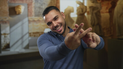 Young hispanic man makes hashtag sign with fingers near classical sculpture in museum gallery hall; joy.