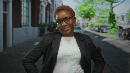 Young african american woman stands with hands on hips in urban street scene framed by buildings and parked cars; confidence empowerment.