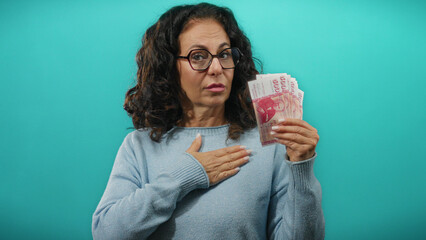 Middle-aged woman holding icelandic krona banknotes while raising hand in oath gesture against a blue wall background.